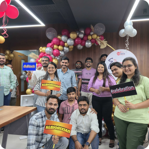 Group of coworkers posing at an office celebration with colorful balloons and festive signs, smiling for the photo.